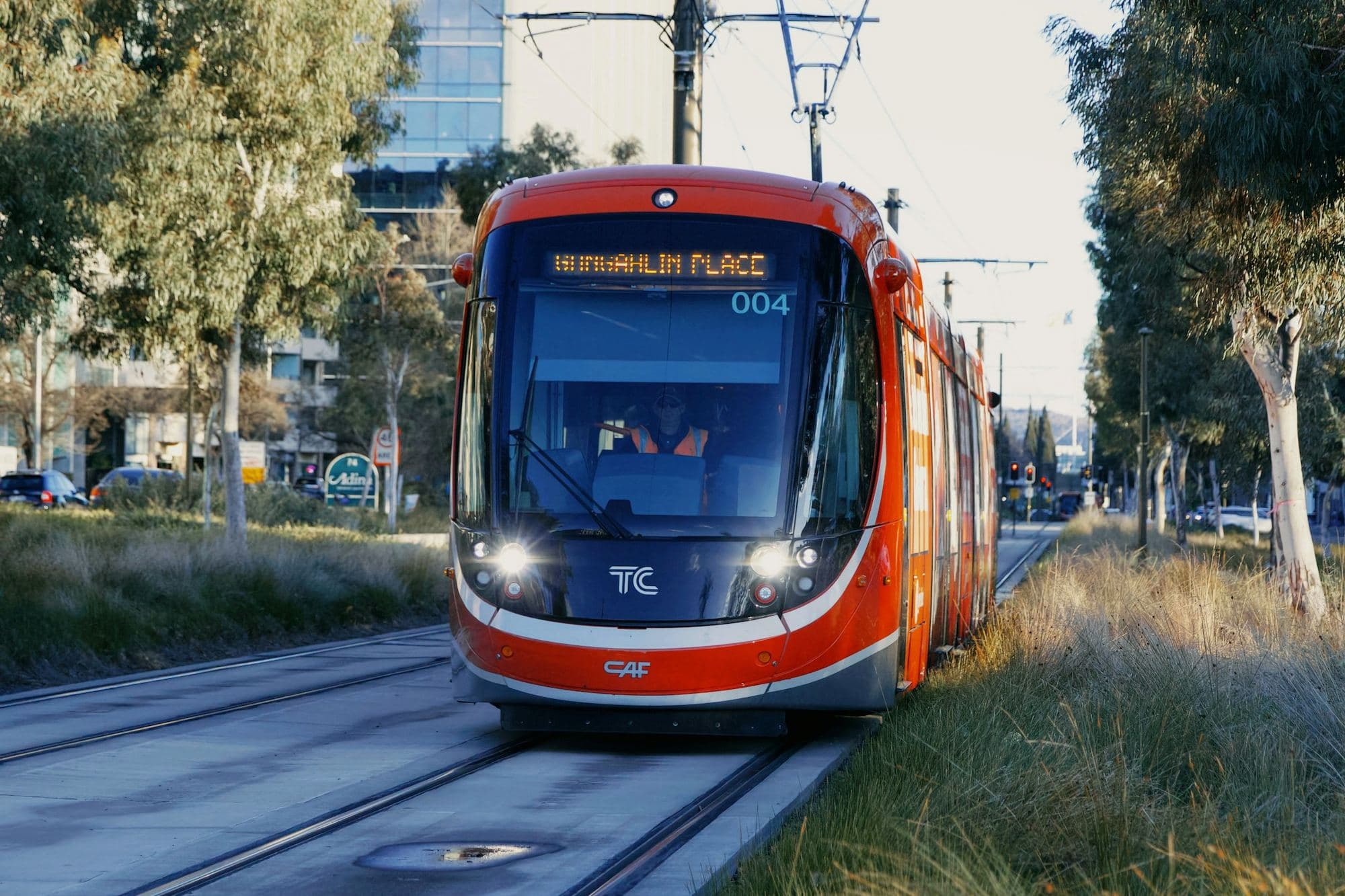 A red Canberra light rail vehicle heading to Gungahlin Place along a tree-lined corridor in soft afternoon light