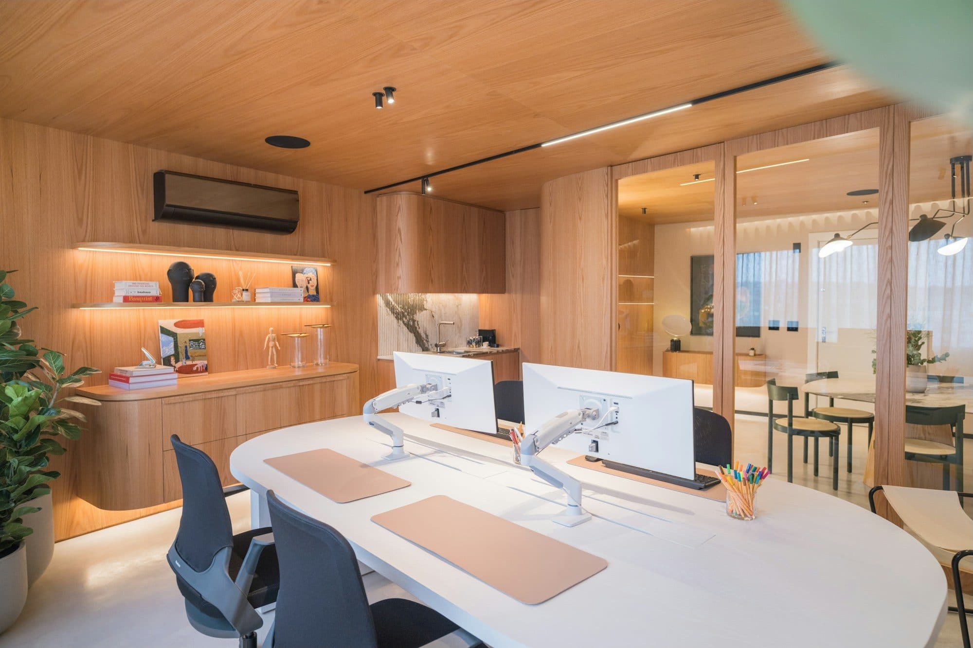 A bright private office with timber joinery, a white desk with two monitors and soft warm lighting
