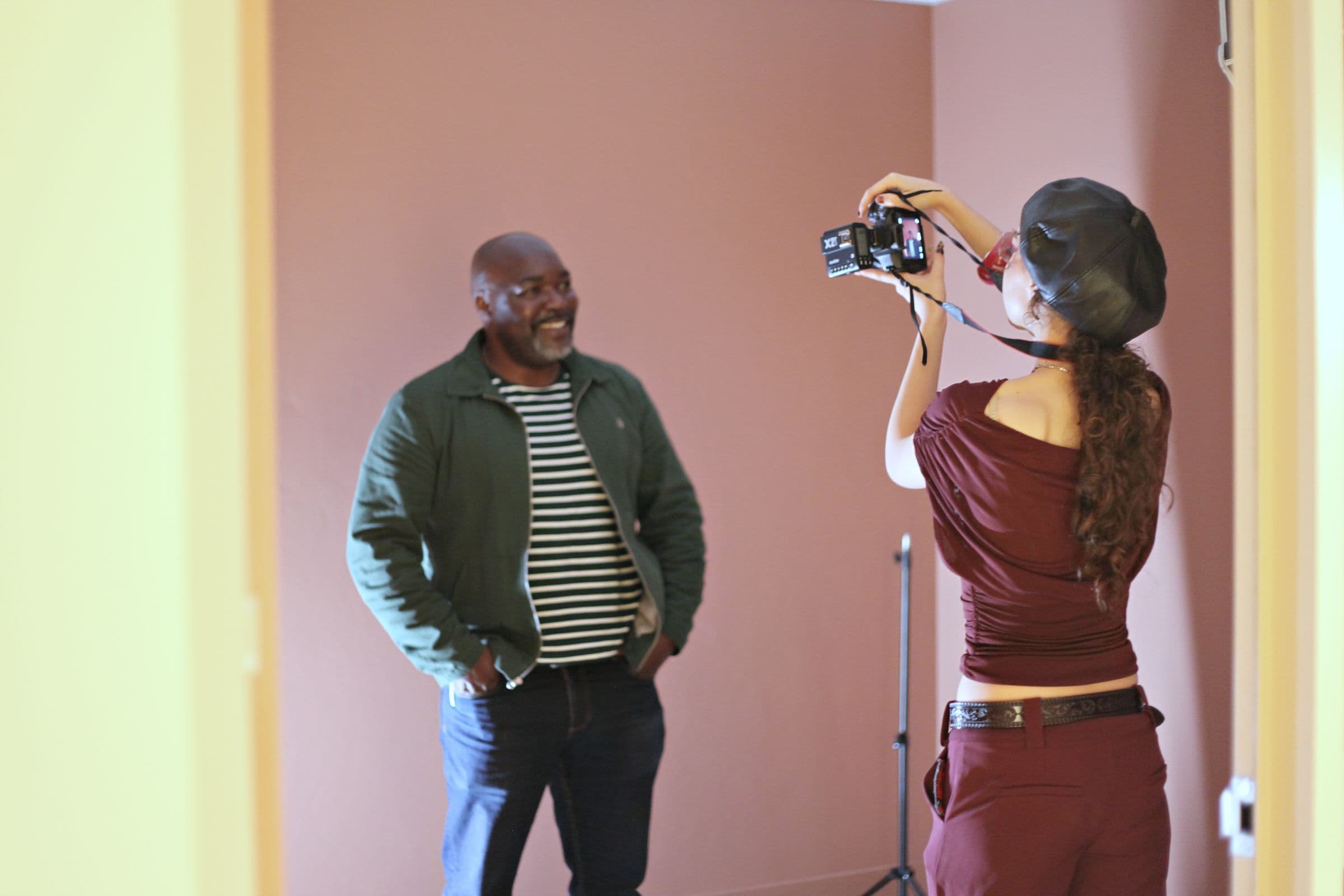 A photographer framing a portrait of a man against a pink wall, with a light stand behind